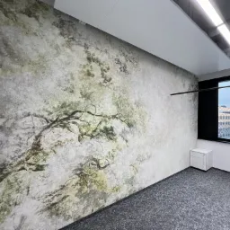 Ściana biura z fototapetą przedstawiającą drzewo, szara wykładzina podłogowa, biała szafka i czarne krzesło biurowe pod oknem.