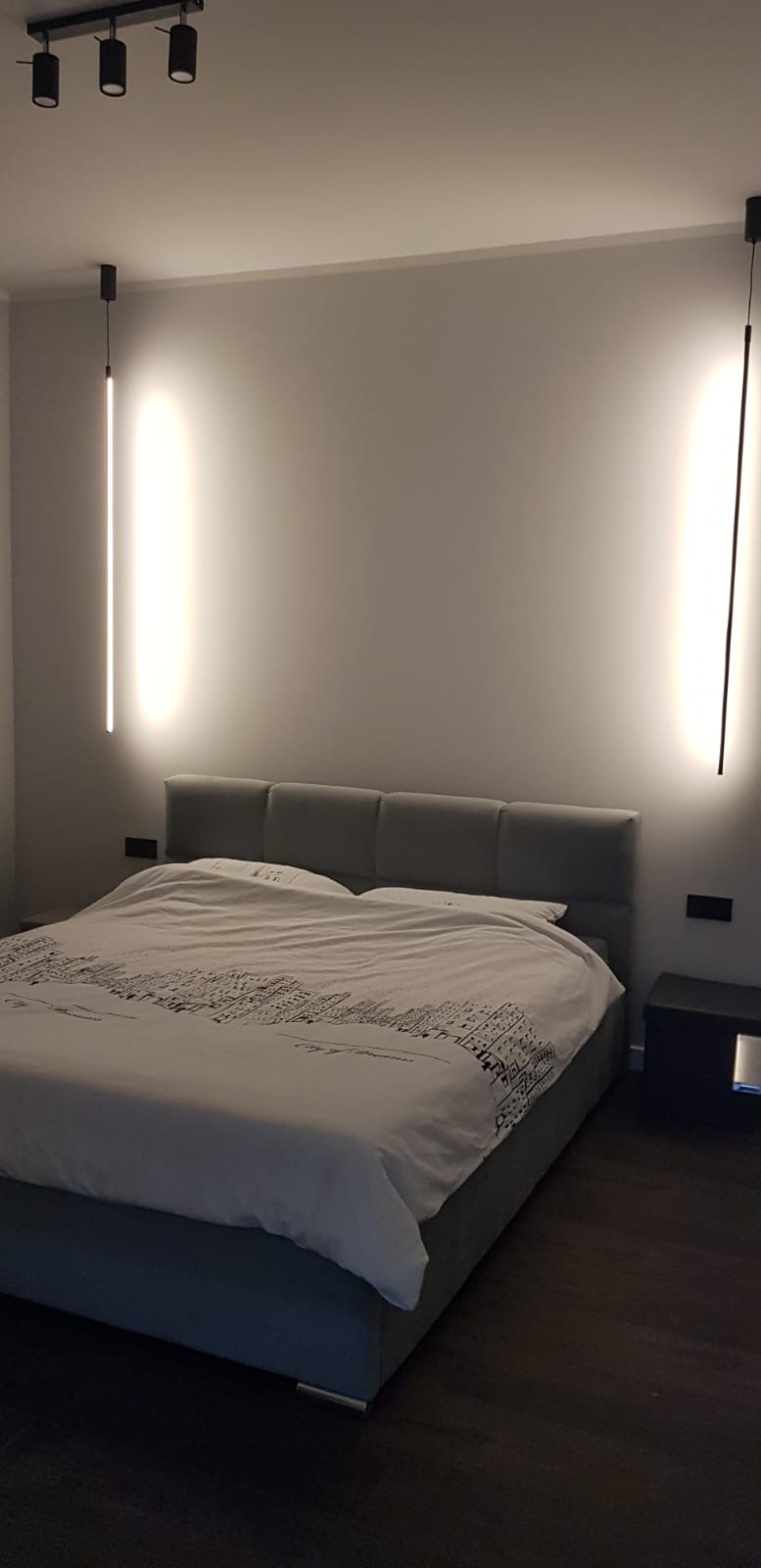 Sypialnia w minimalistycznym stylu z szarym łóżkiem kontynentalnym, pościelą w miejski wzór i pionowymi lampami LED po bokach.