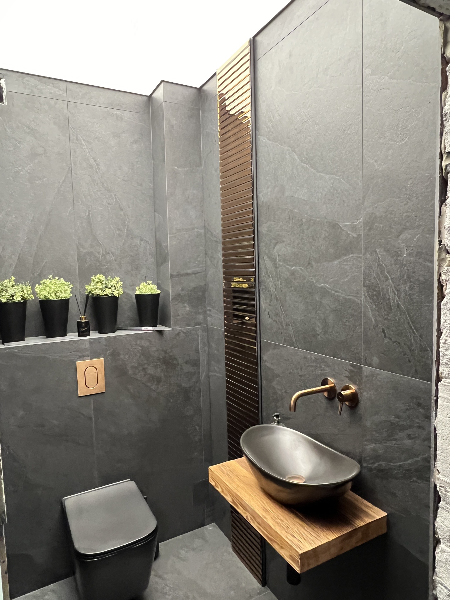 Nowoczesna łazienka w odcieniach szarości z umywalką nablatową w kształcie misy, drewnianym blatem, złotą armaturą i czarną toaletą wiszącą; na półce wnękowej cztery doniczki z zielonymi roślinami...