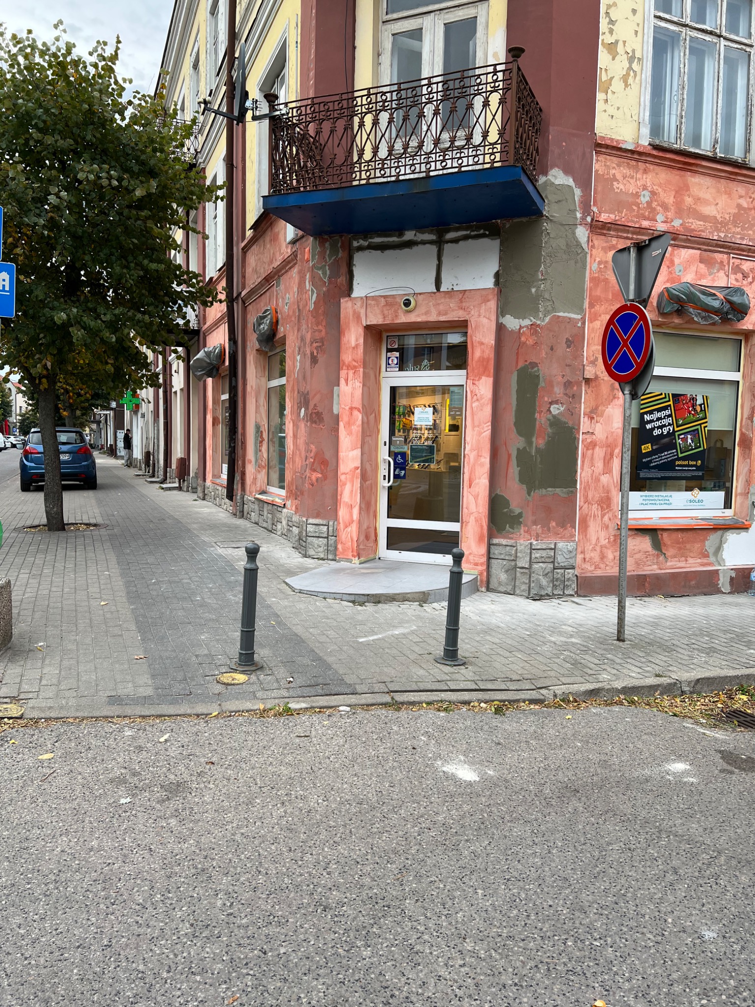 Fasada budynku w trakcie renowacji z widocznymi śladami starej farby i świeżo nałożonym tynkiem, wejście z udogodnieniami dla niepełnosprawnych, znak zakazu parkowania w tle.
