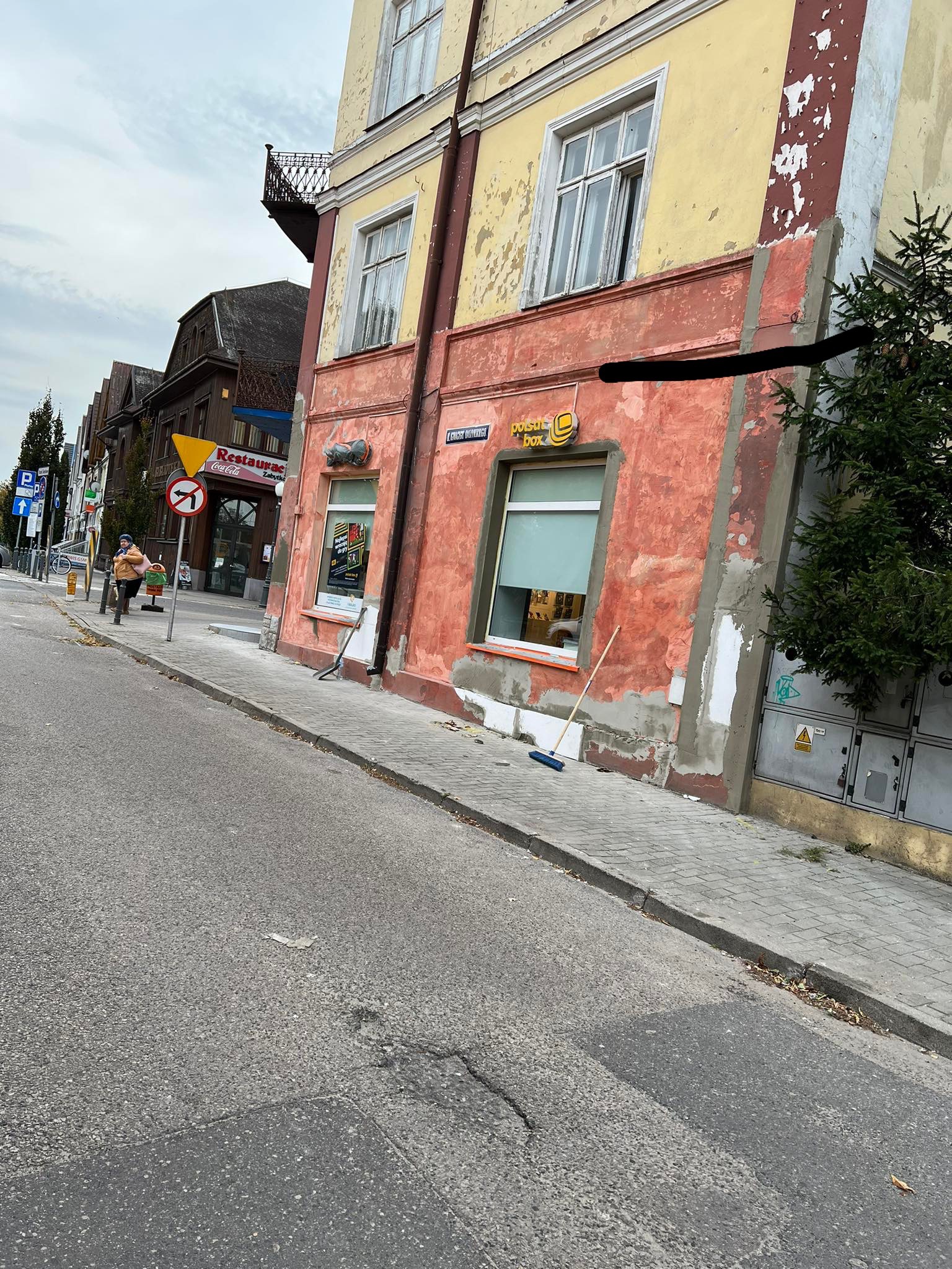 Elewacja budynku w trakcie renowacji z widocznymi śladami po usuniętej farbie, częściowo pomalowana na czerwono i żółto, z oknami i szyldem 'Polska Box', obok stoi miotła.