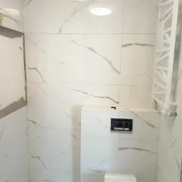 Nowoczesna toaleta wisząca z czarnym przyciskiem spłukującym, otoczona białymi płytkami imitującymi marmur, z grzejnikiem drabinkowym owiniętym folią ochronną po prawej stronie i wentylatorem...