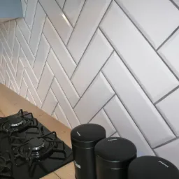 Białe, prostokątne płytki ułożone w jodełkę na ścianie nad blatem kuchennym z czarną płytą gazową i zestawem czarnych pojemników na przyprawy.