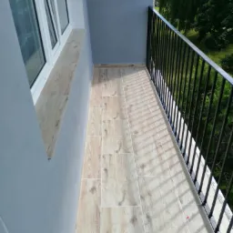 Wykończony balkon z szarą ścianą, białą ramą okienną i podłogą wyłożoną panelami imitującymi drewno, czarna metalowa balustrada z widokiem na zieleń.