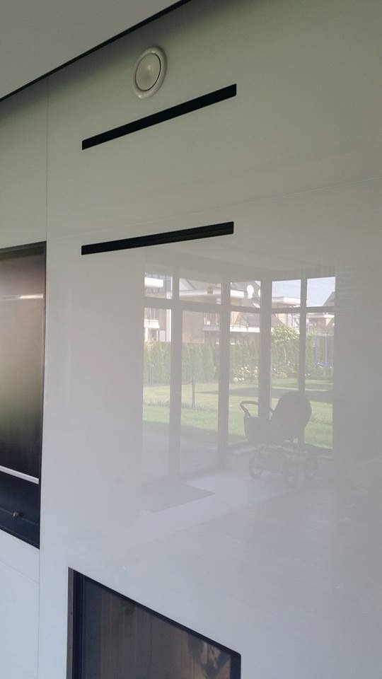 Błyszcząca, biała ściana z czarnymi, poziomymi akcentami i okrągłym nawiewnikiem wentylacyjnym, odbijająca widok okna z ogrodem i wózkiem dziecięcym w tle.