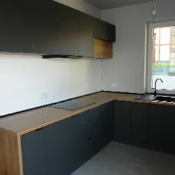 Nowoczesna kuchnia z grafitowymi szafkami i drewnianym blatem, minimalistyczny design, widoczne gniazdka elektryczne i wystające kable instalacji elektrycznej.