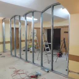 Stelaż z profili metalowych tworzący łukowate przejście, widoczne drabiny, narzędzia i laserowy niwelator w pomieszczeniu w trakcie prac wykończeniowych.