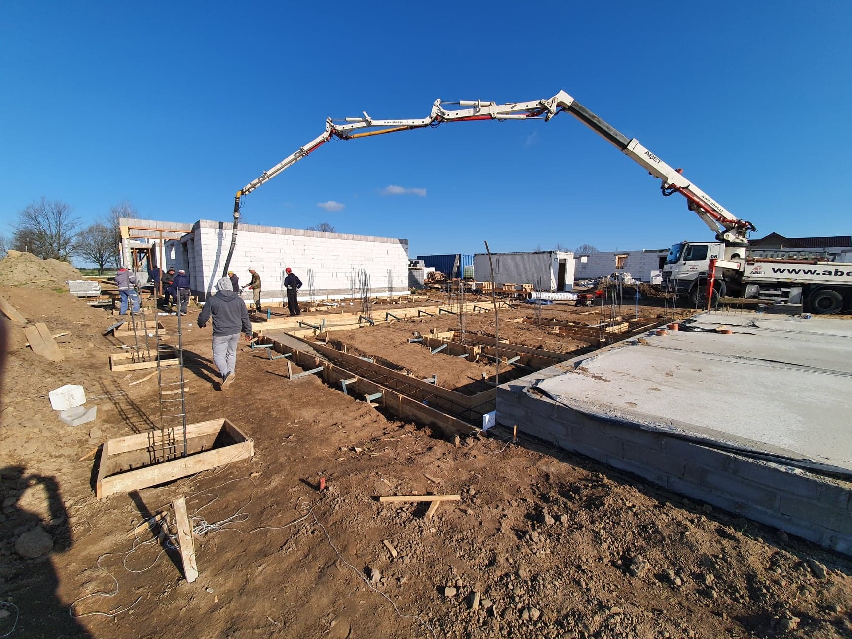 Wylewanie betonu do szalunków zbrojeniowych pod fundamenty domu jednorodzinnego, widoczny betonowóz z pompą oraz ekipa budowlana w trakcie prac.
