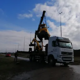Załadunek koparki na lawetę ciężarową Volvo z logo Gkmediawawa, w tle pochmurne niebo i czerwony samochód osobowy.