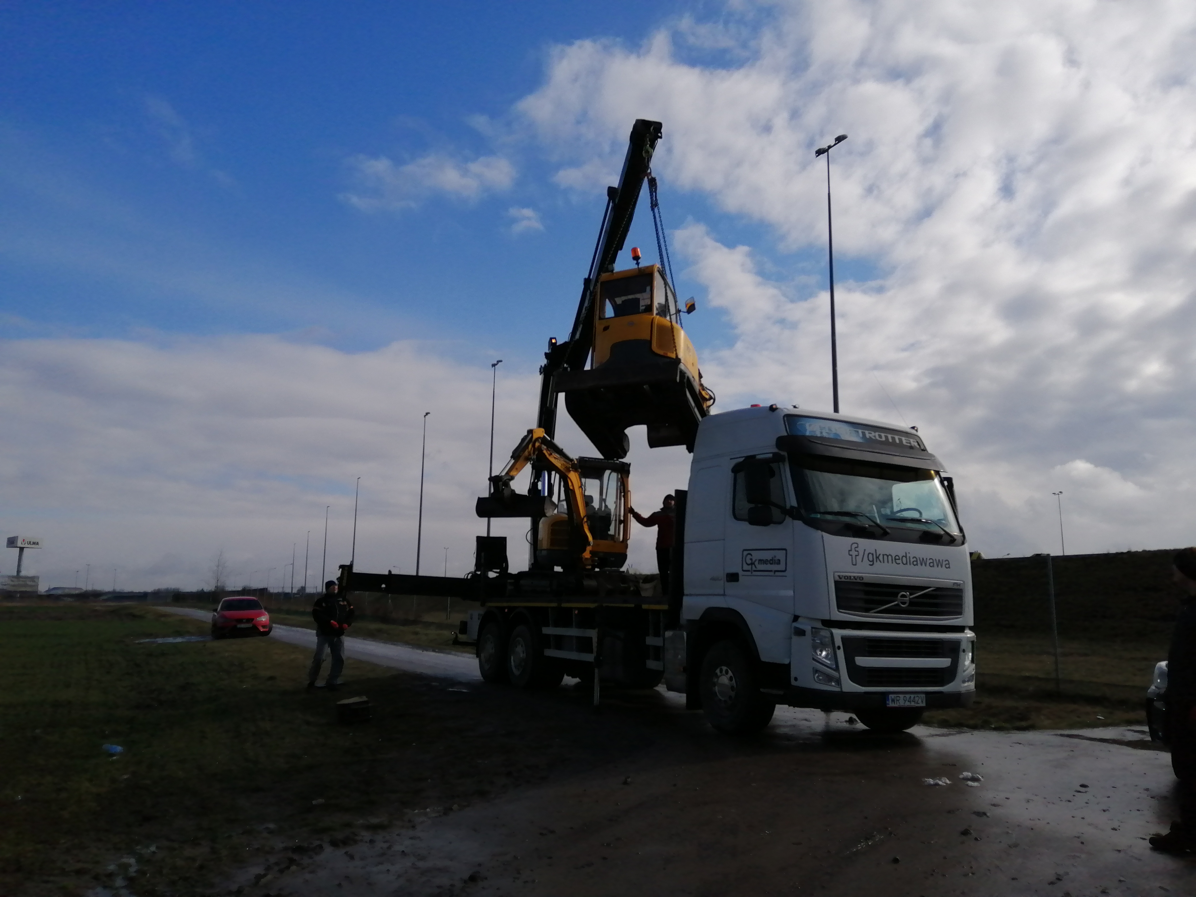 Załadunek koparki na lawetę ciężarową Volvo z logo Gkmediawawa, w tle pochmurne niebo i czerwony samochód osobowy.
