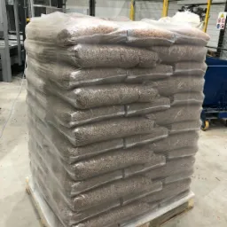 Ułożone na palecie, owinięte folią worki z brązowym granulatem, prawdopodobnie paliwem ekologicznym, w tle hala produkcyjna z maszynami.