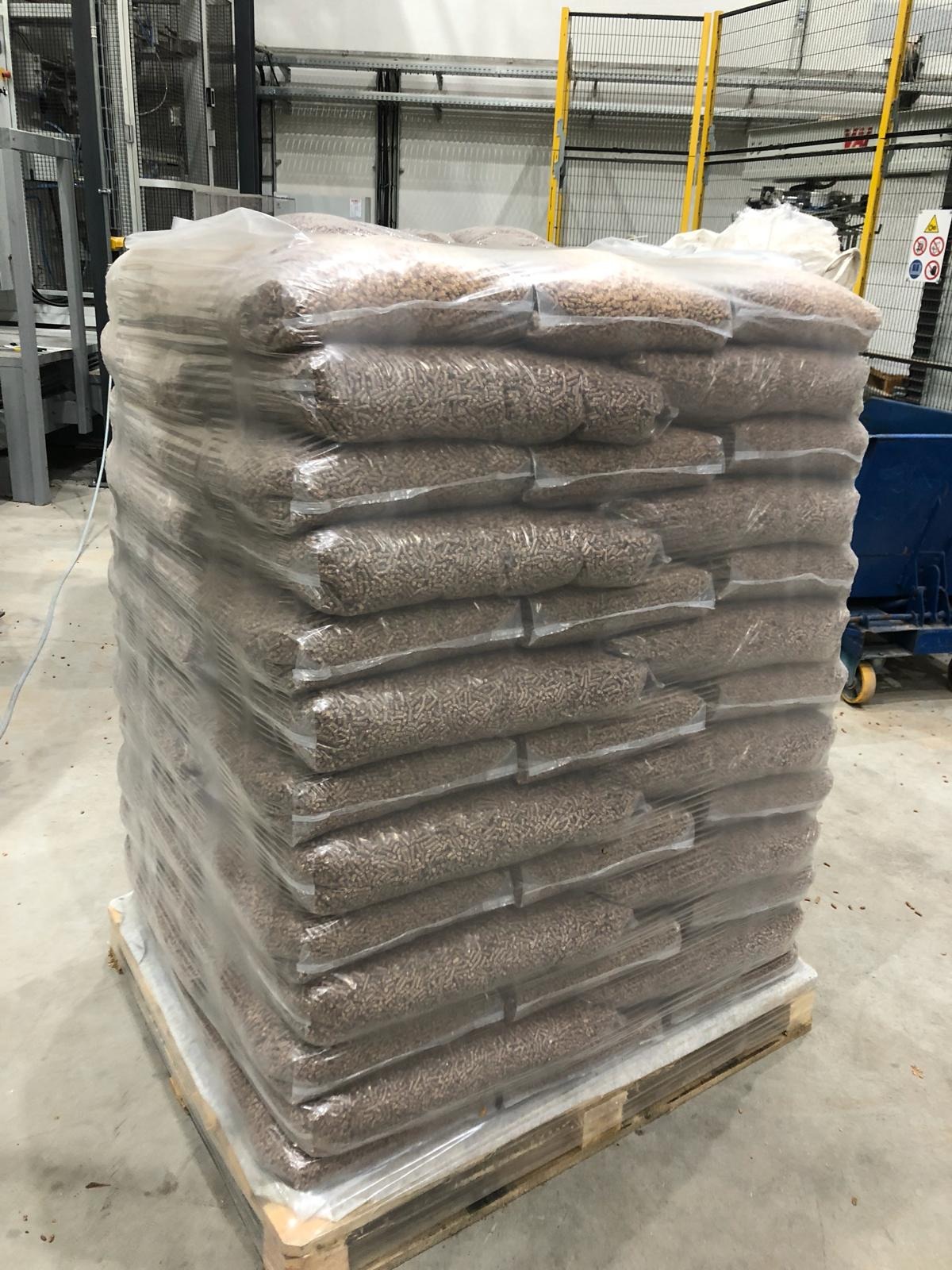 Ułożone na palecie, owinięte folią worki z brązowym granulatem, prawdopodobnie paliwem ekologicznym, w tle hala produkcyjna z maszynami.