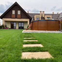 Utrzymany trawnik z kamienną ścieżką prowadzącą do drewnianego basenu naziemnego na tle domu z brązowym dachem.