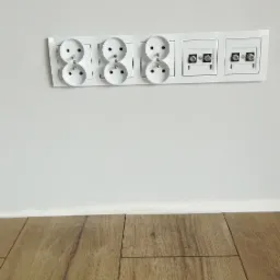 Biała listwa z trzema gniazdkami elektrycznymi typu francuskiego i dwoma gniazdami antenowymi na białej ścianie, widok z dołu, jasne panele podłogowe.