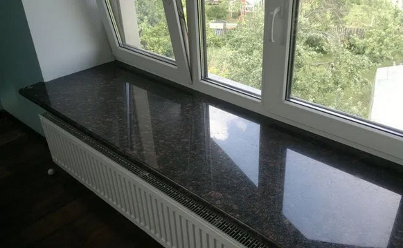 Błyszczący, ciemnobrązowy parapet z konglomeratu, zamontowany pod białym oknem, z widocznym grzejnikiem żeberkowym i fragmentem drewnianej podłogi.