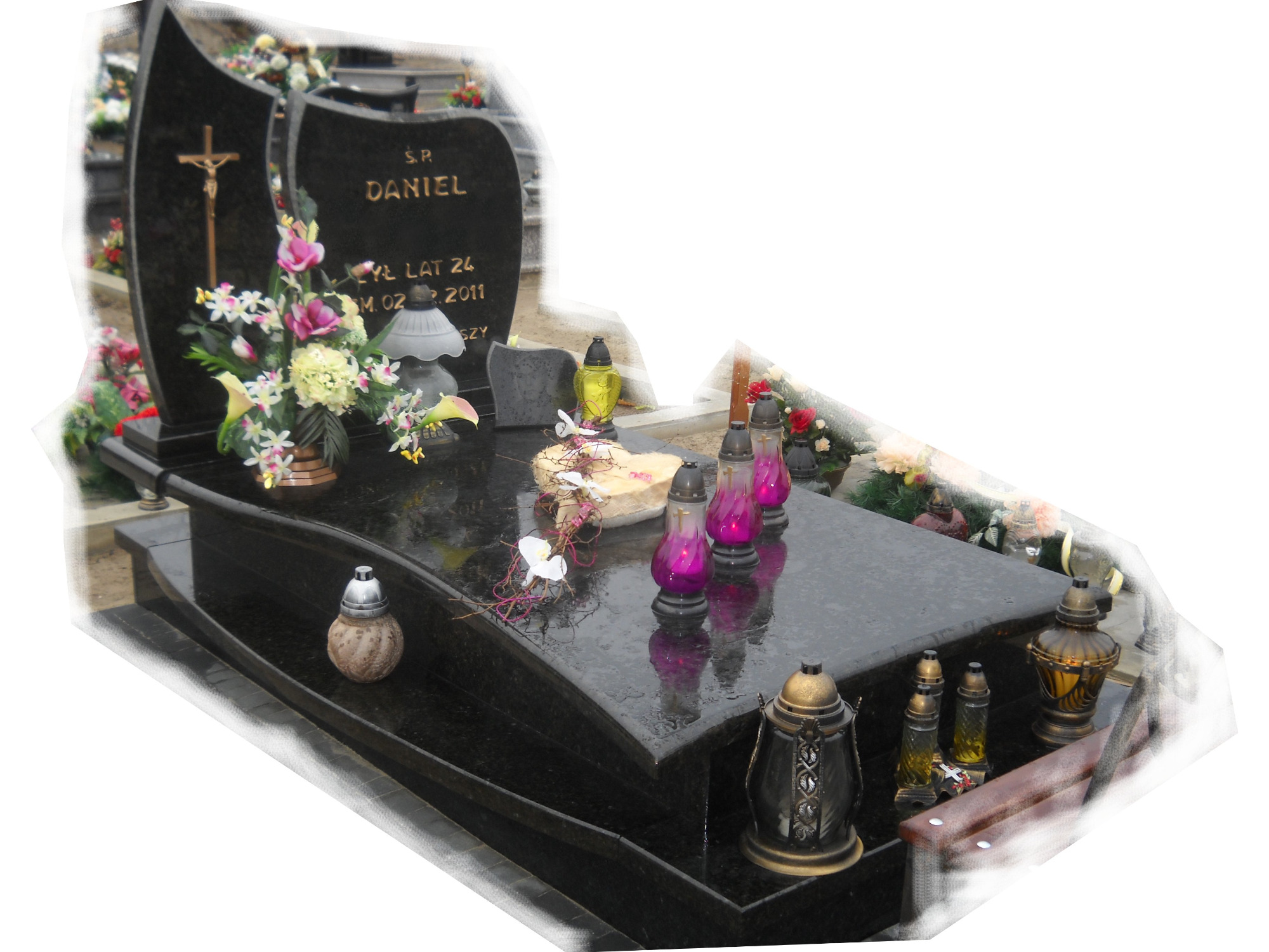 Czarny, polerowany pomnik z kwiatami i zniczami, z wygrawerowanym imieniem i nazwiskiem 'S.P. Daniel', w tle inne nagrobki.