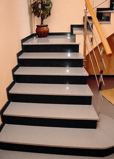 Wewnętrzne schody z szarymi stopniami i czarnymi podstopnicami, wykończone stalową balustradą z drewnianym poręczą, oświetlone punktowo na podeście, z rośliną doniczkową w tle.