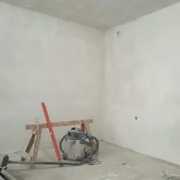 Pokój w trakcie prac wykończeniowych, ściany i sufit pokryte świeżą warstwą, widoczny odkurzacz przemysłowy, poziomica i podpory na podłodze.
