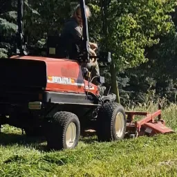 Operator na czerwonym traktorze Shibaura kosi wysoką trawę na tle drzew. Sprzęt w akcji, widok z boku, zieleń i maszyna w ruchu.