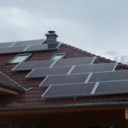 Panele słoneczne zamontowane na dachu z czerwonej dachówki, widoczne okna dachowe i komin, pochmurne niebo w tle.