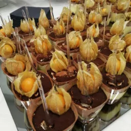 Deser tiramisu w małych porcjach, udekorowany miechunką peruwiańską i mini łyżeczką, ustawiony na srebrnej tacy, widoczne inne przekąski w tle.