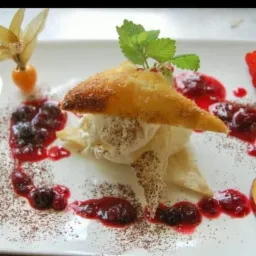 Elegancko podany deser na białym talerzu: tr&oacute;jkątny pierożek z ciasta francuskiego na kremie, udekorowany sosem z owoc&oacute;w leśnych, miechunką i listkiem mięty, posypany kakao.