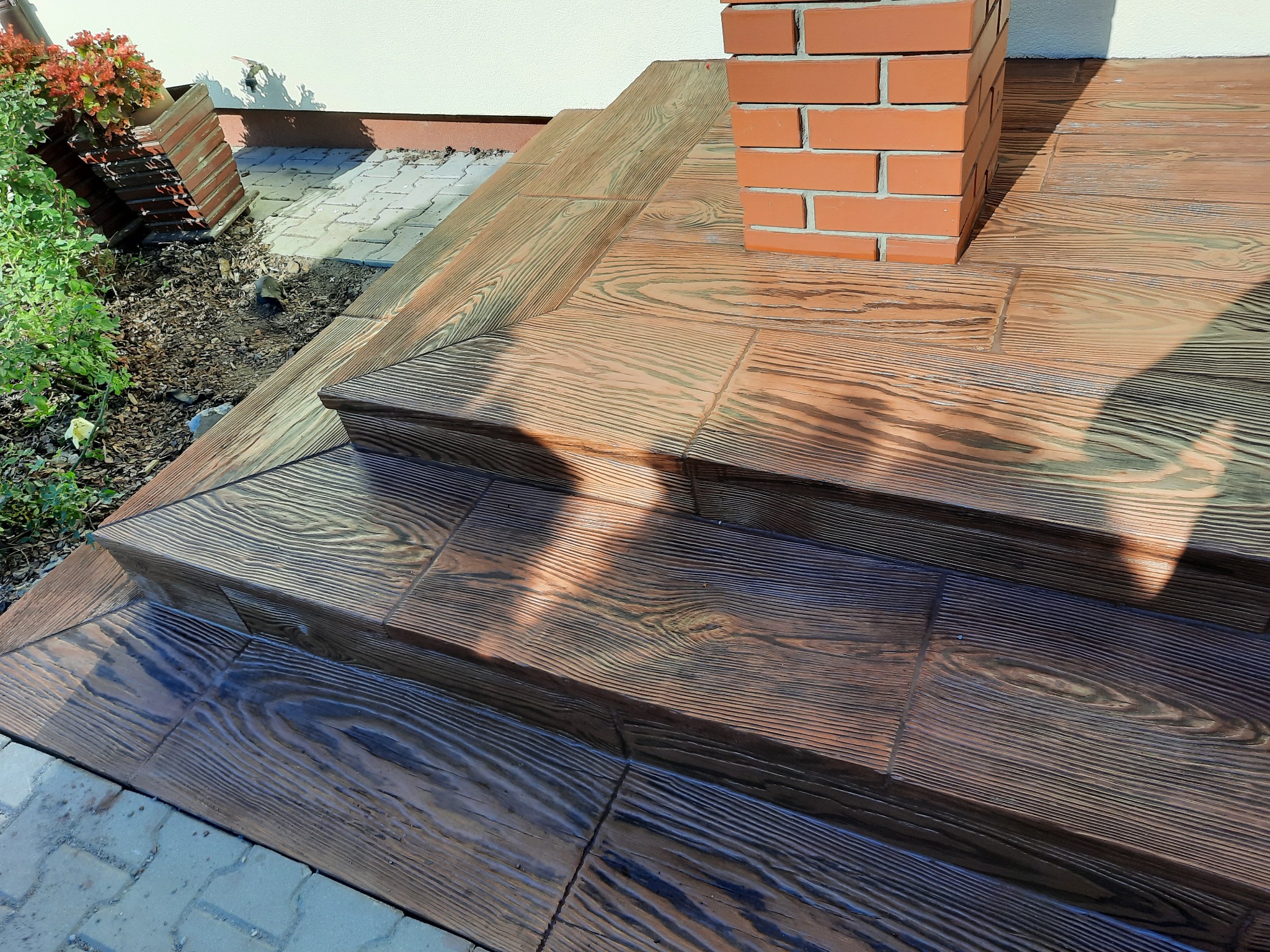 Schody zewnętrzne wyłożone brązową kostką z fakturą drewna, zakończone ceglanym słupkiem przy ścianie budynku.