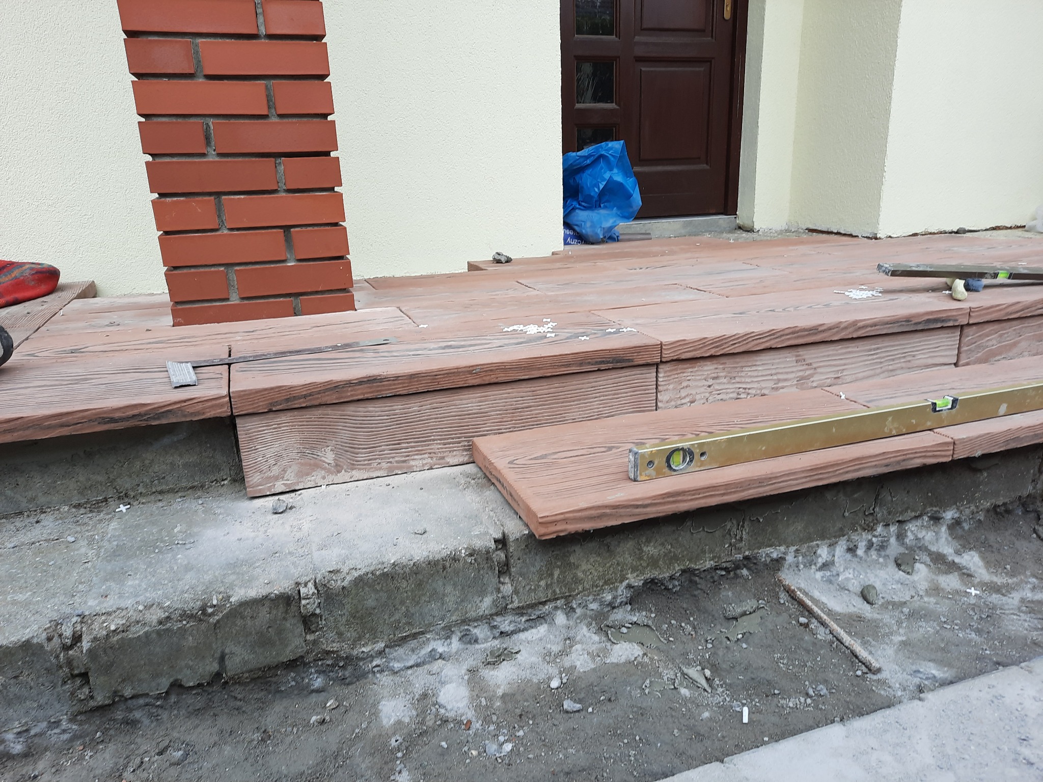 Montaż drewnopodobnych stopni wejściowych do domu, widoczne poziomica, fragment ceglanego filaru i niebieski worek przy drzwiach.