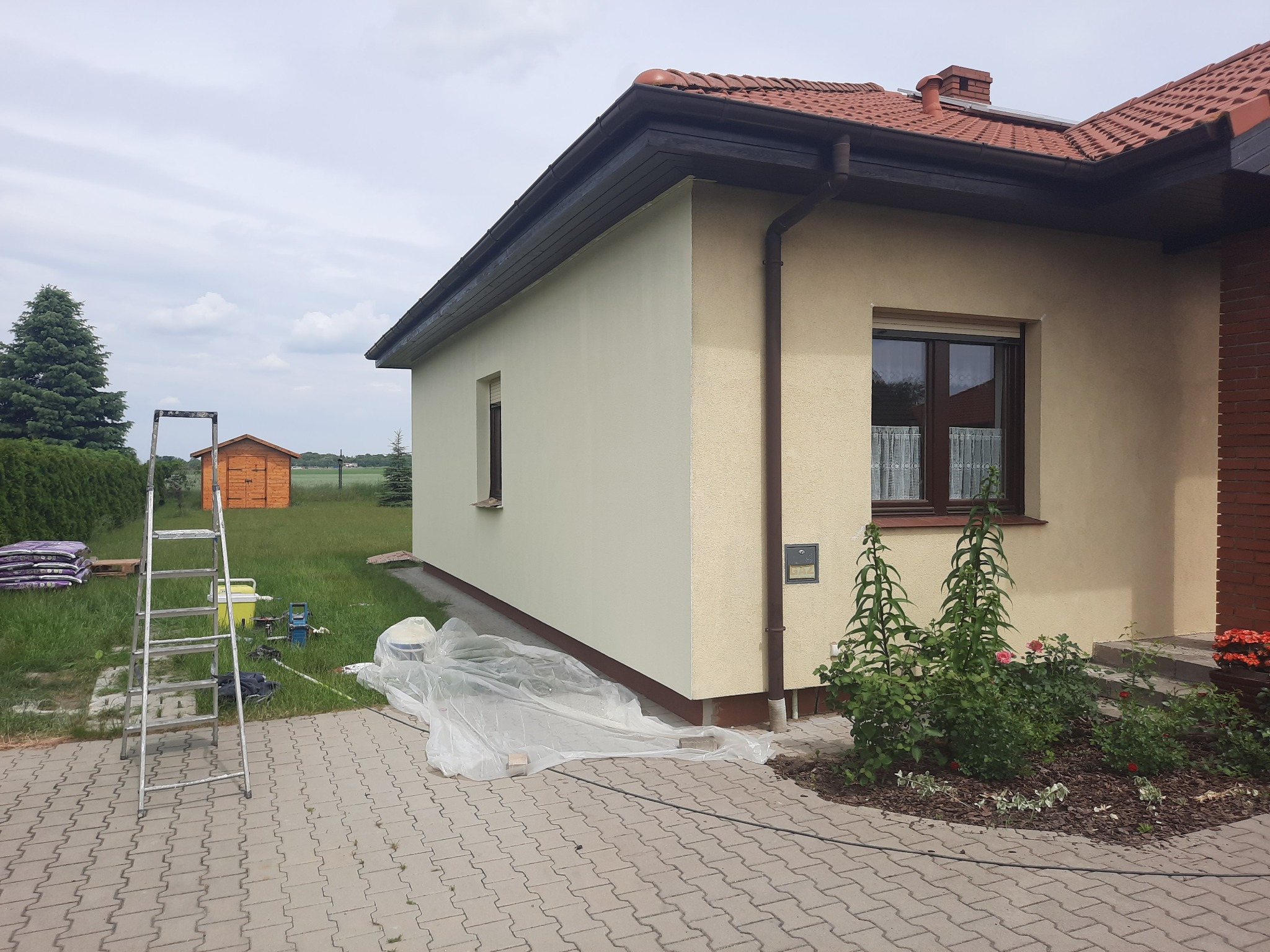 Dom jednorodzinny po częściowym myciu elewacji; widoczna drabina, worki z materiałami, zabezpieczona folią część ściany, świeżo umyta jasna elewacja kontrastuje z fragmentem elewacji w kolorze...