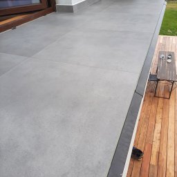 FLIZ-DEM - Wykończony balkon z szarą, dużą płytką gresową na podłodze, drewniany taras z małym stolikiem i widokiem na zielone wzgórza w tle.