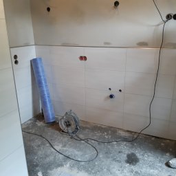 FLIZ-DEM - Białe płytki ścienne w trakcie montażu, widoczne puszki elektryczne, fragment instalacji wodnej, zwinięta folia hydroizolacyjna oraz przedłużacz na podłodze podczas prac wykończeniowych.