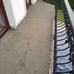FLIZ-DEM - Wyrównana, świeża wylewka na balkonie w Jordanowie, przygotowana pod dalsze prace wykończeniowe, widoczne ozdobne, czarne balustrady i okna balkonowe.