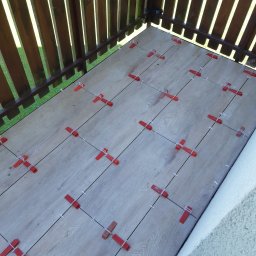FLIZ-DEM - Balkon w trakcie układania płytek imitujących drewno, z użyciem czerwonych klipsów poziomujących i białych krzyżyków dystansowych, widok z góry na drewnianą balustradę i fragment elewacji.