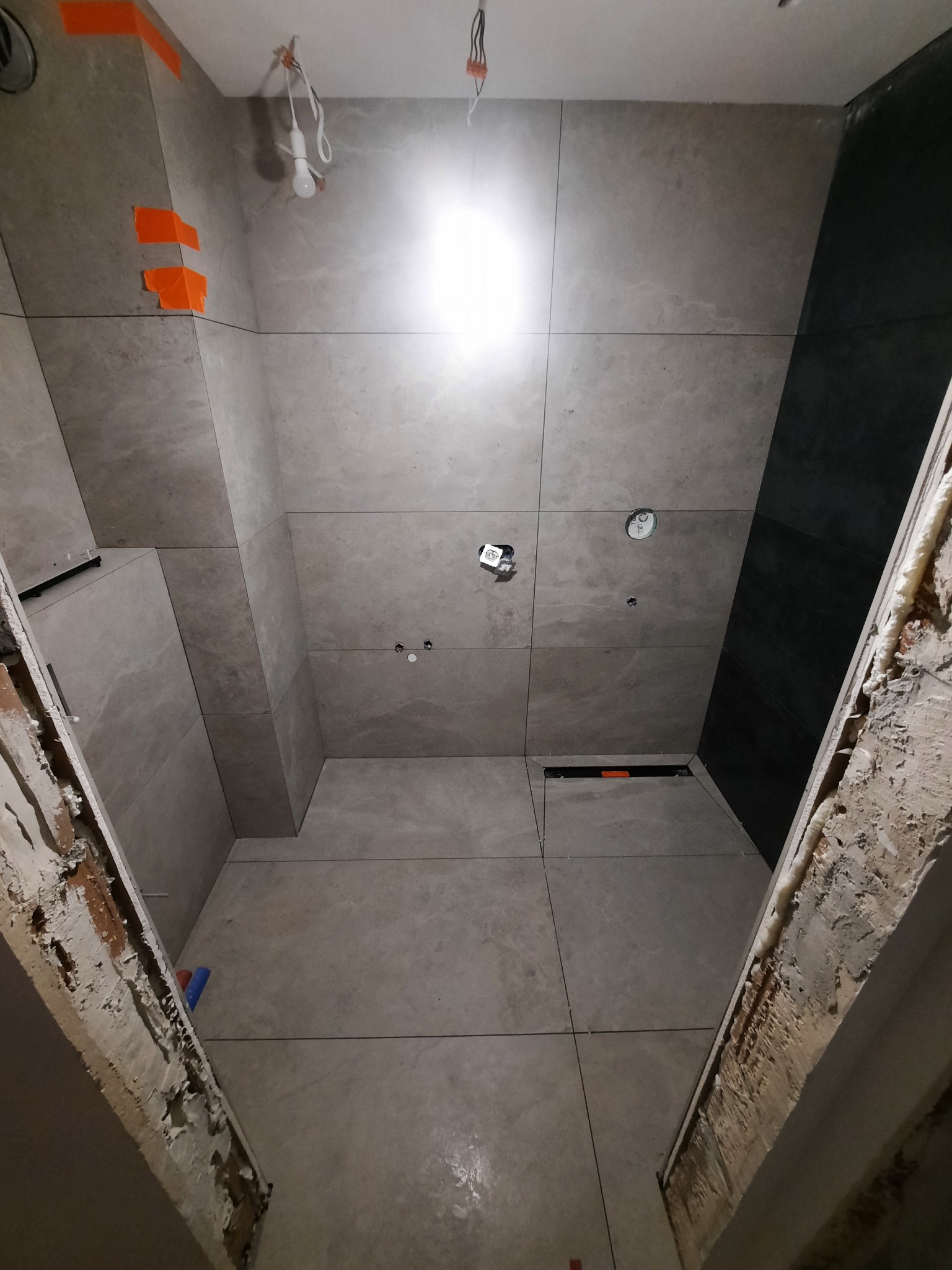 Wnętrze łazienki w trakcie wykańczania; widoczne szare płytki na ścianach i podłodze, fragment czarnej ściany, niedokończone obramowanie drzwi z pianką montażową oraz prowizoryczne oświetlenie.