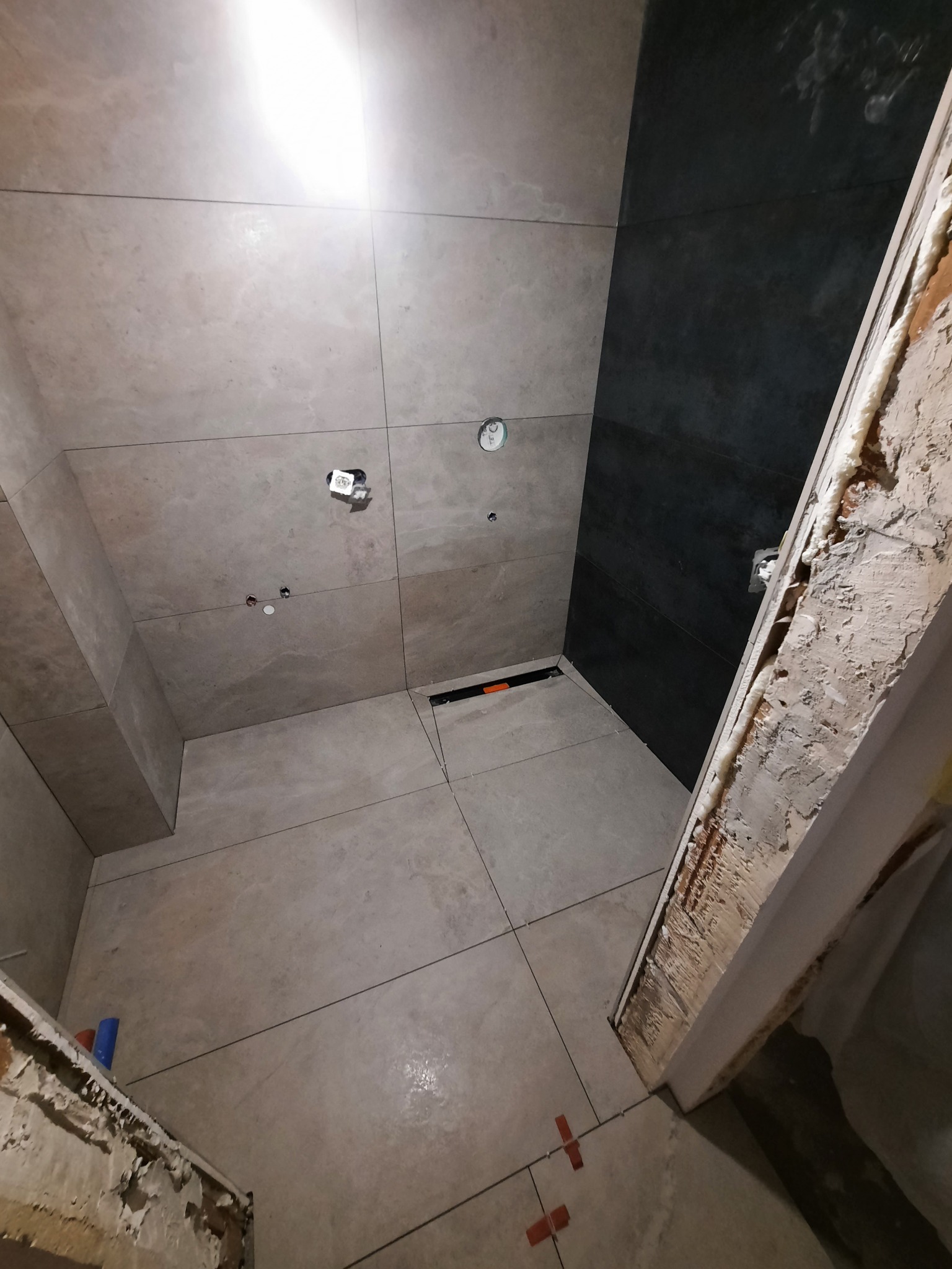 Wnętrze łazienki w trakcie remontu, widoczne szare płytki na ścianach i podłodze, czarny fragment ściany, wystające instalacje oraz fragment futryny z pianką montażową.