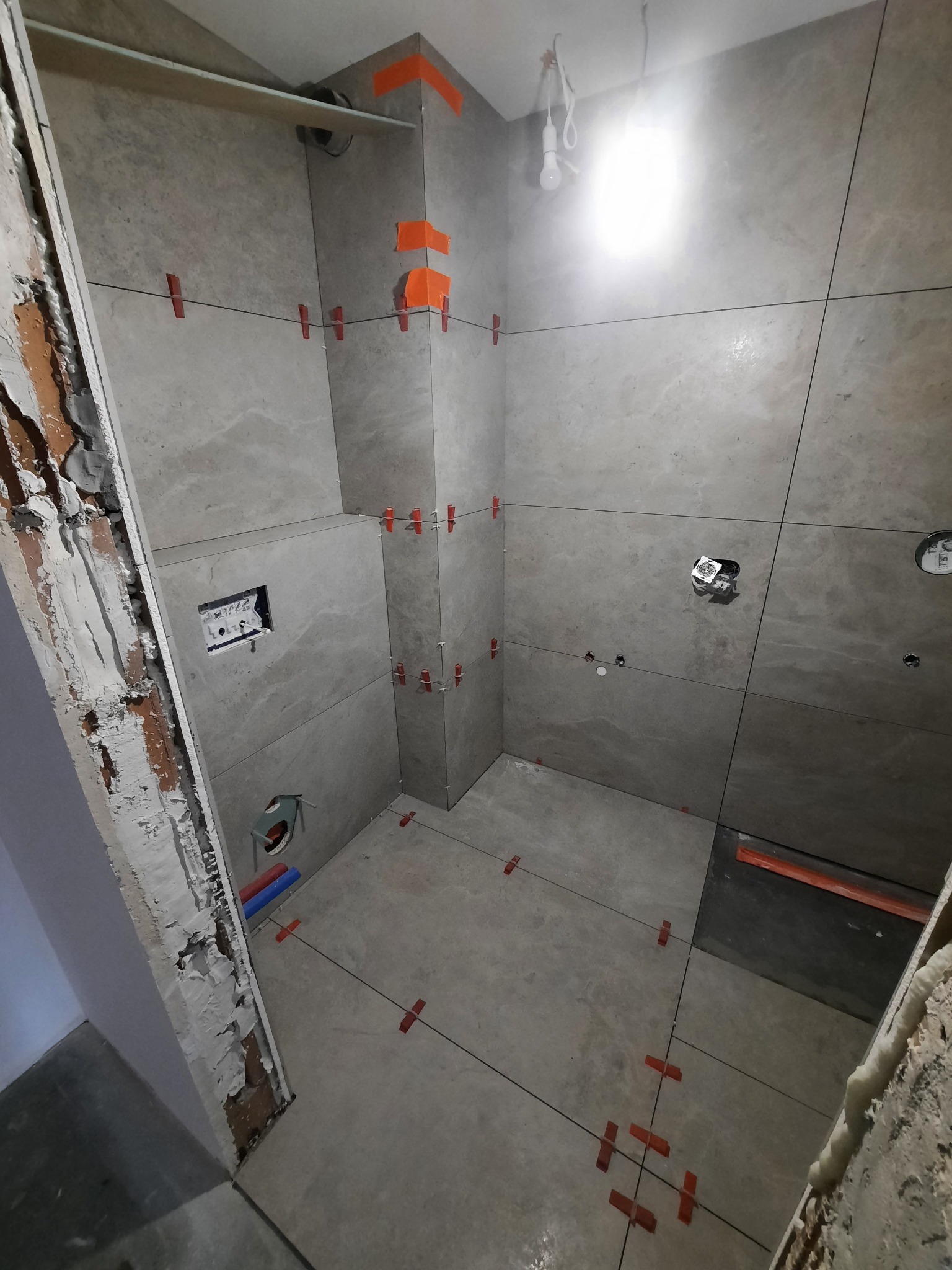 Łazienka w trakcie remontu: szare, duże płytki na ścianach z pomarańczowymi krzyżykami dystansowymi, widoczne instalacje hydrauliczne i elektryczne, surowa ściana z odsłoniętą cegłą.