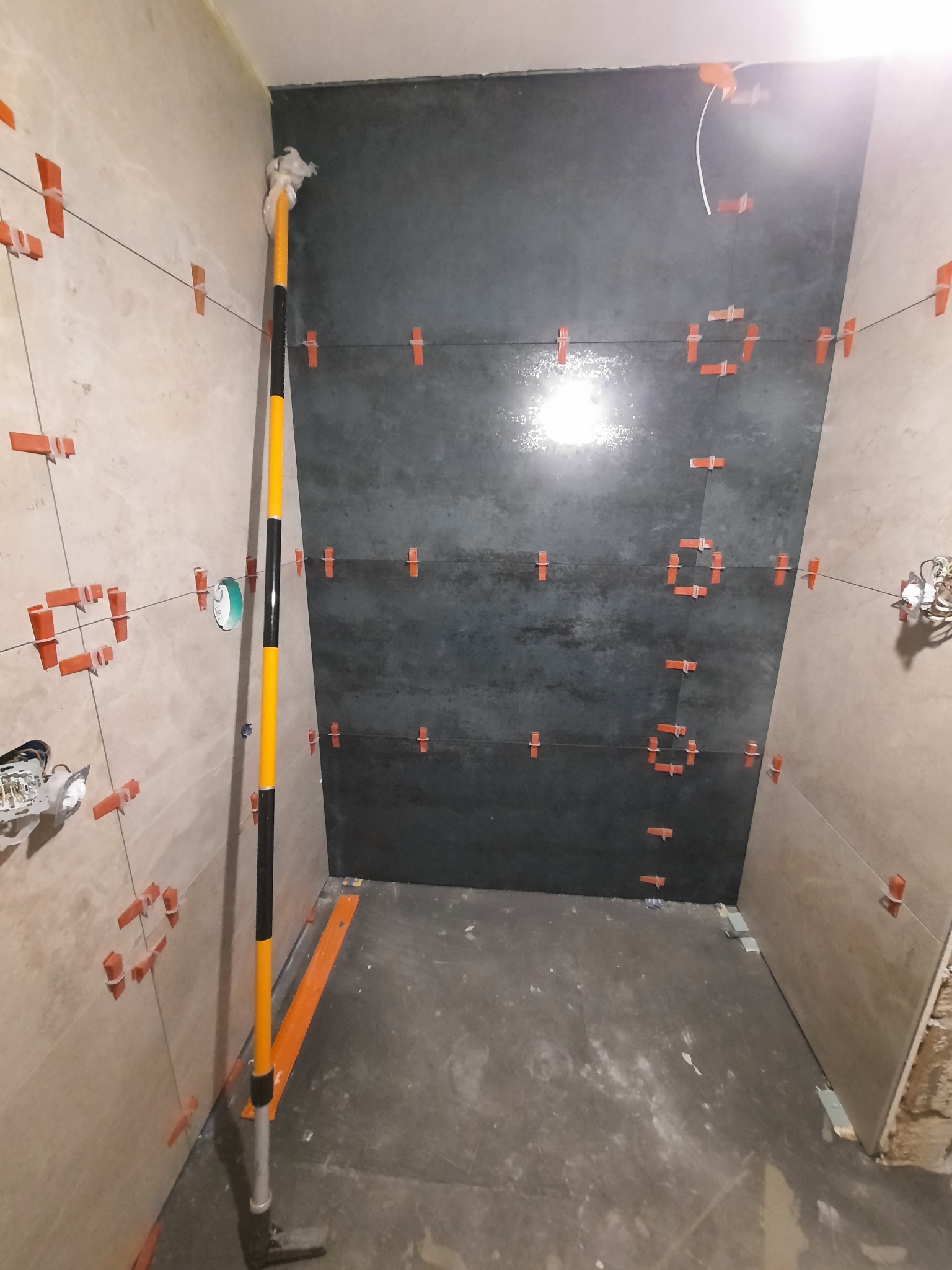 Ściany w trakcie układania płytek w łazience, widoczne płytki w odcieniach szarości i beżu, pomarańczowe klipsy poziomujące, sznurki, poziomica, oraz surowy beton na podłodze.