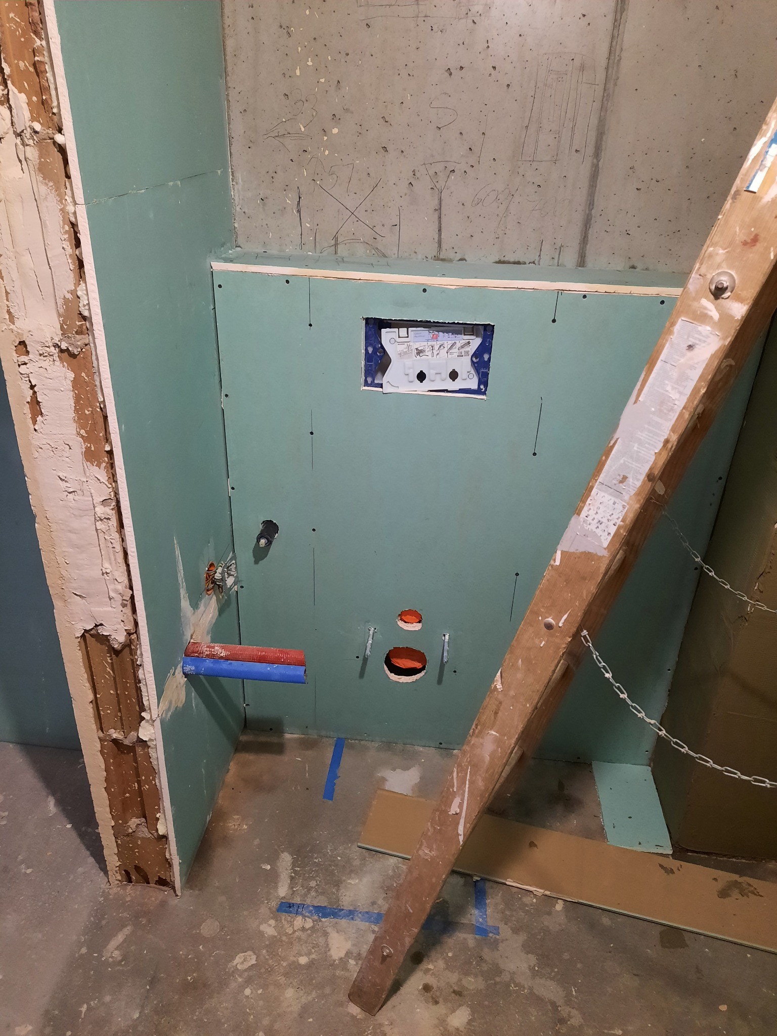 Wnętrze w trakcie remontu: ściana z płyt kartonowo-gipsowych w kolorze zielonym, widoczne instalacje hydrauliczne z rurami w kolorze niebieskim i czerwonym, otwory w ścianie, fragment drabiny...