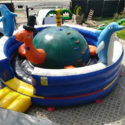 Kolorowy, dmuchany plac zabaw z elementami w kształcie delfinów i dinozaura w cylindrze, ustawiony na betonowej kostce brukowej, z widocznym trampoliną i ogrodzeniem w tle.