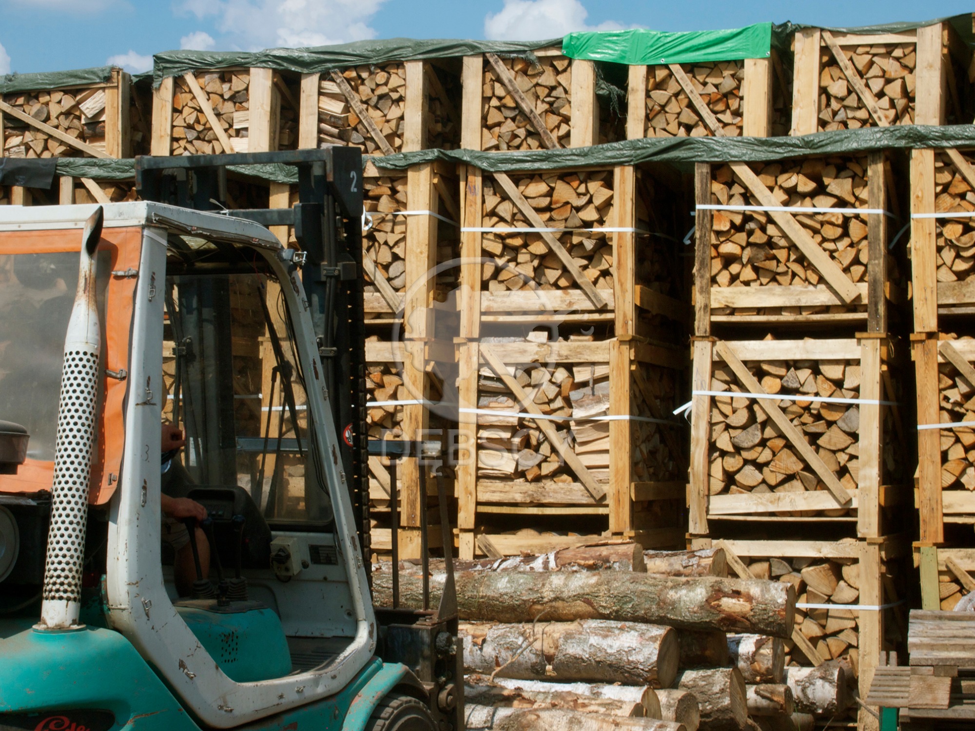 Wózek widłowy przed paletami z drewnem opałowym, część palet przykryta zieloną plandeką.