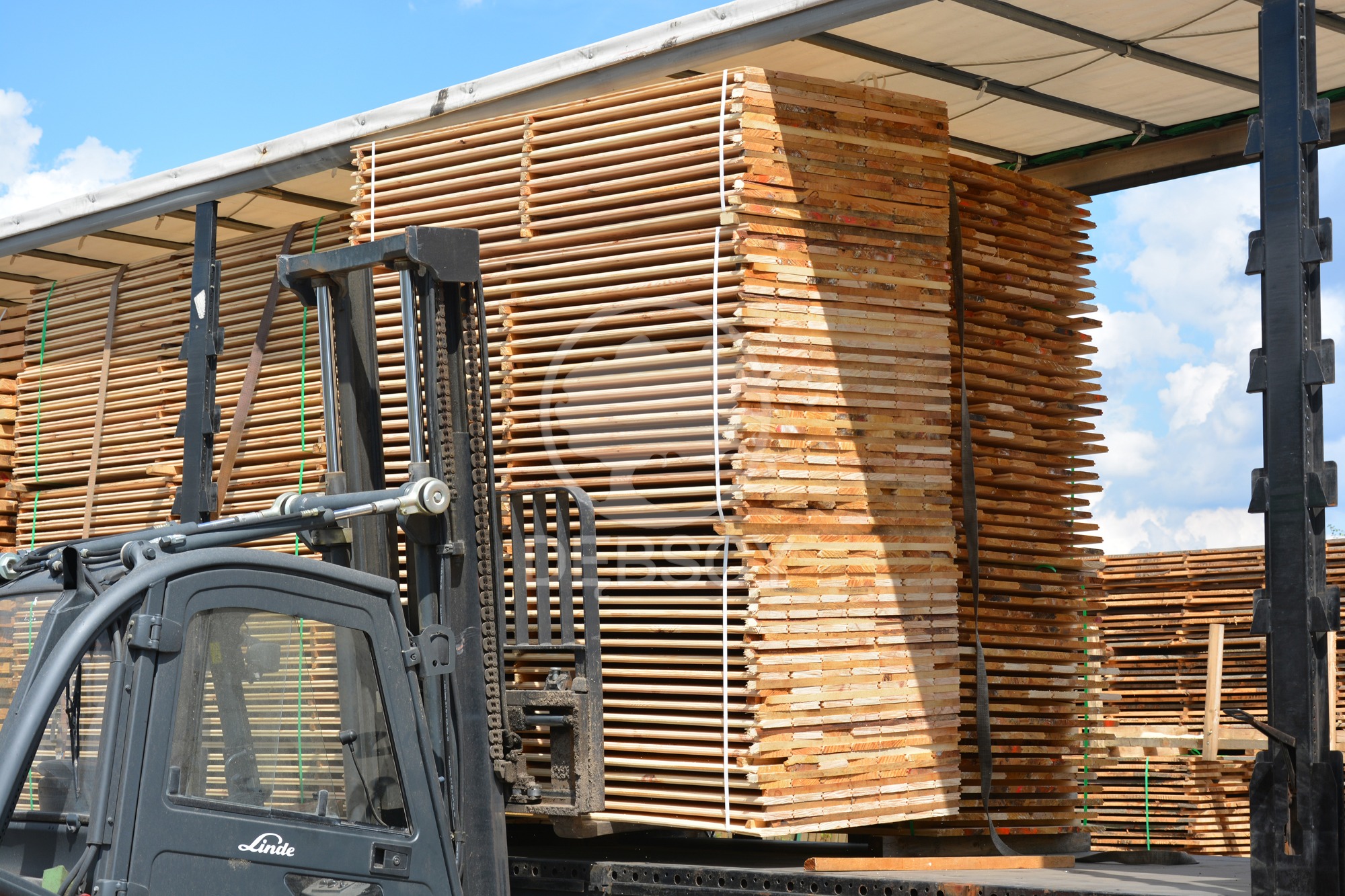Wózek widłowy Linde transportuje palety z jasnego drewna na naczepie, widoczne niebo z chmurami w tle.