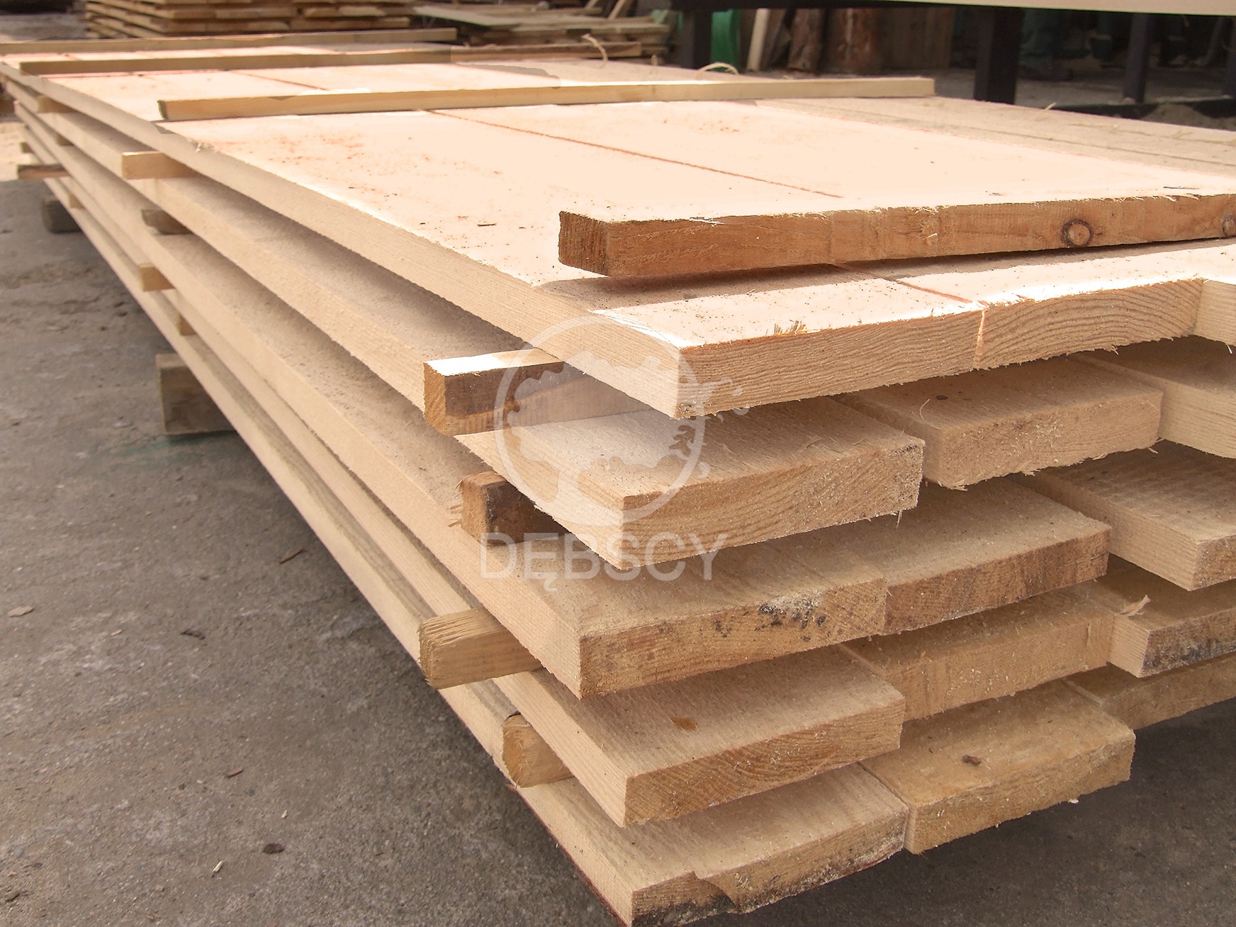 Stos jasnej, świeżo przetartej tarcicy budowlanej ułożonej warstwami na przekładkach, widoczne słoje drewna i delikatne zabrudzenia.