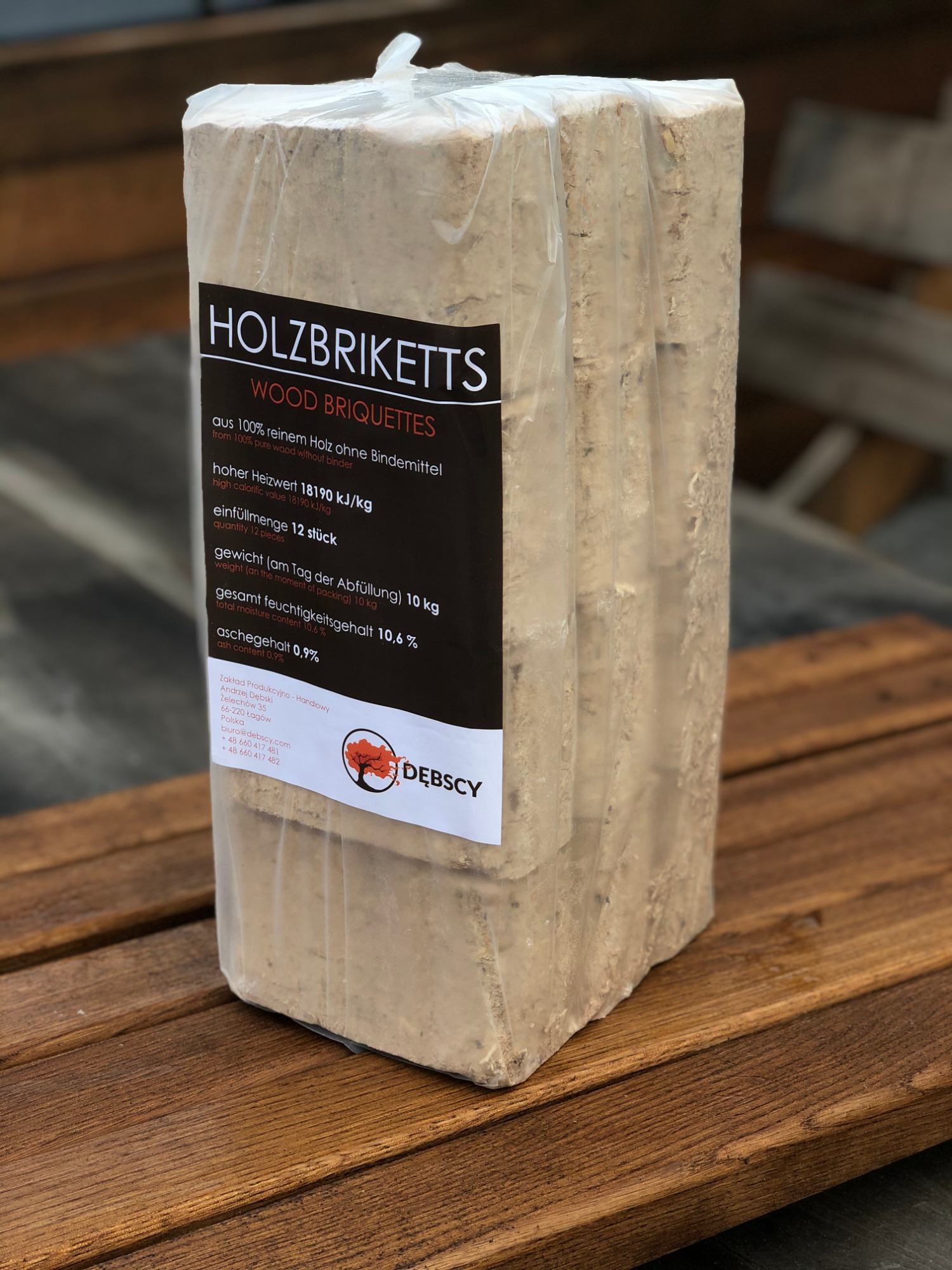Pakiet brykietów drzewnych owinięty w folię, z etykietą informacyjną producenta, leżący na drewnianym blacie z widoczną strukturą drewna.
