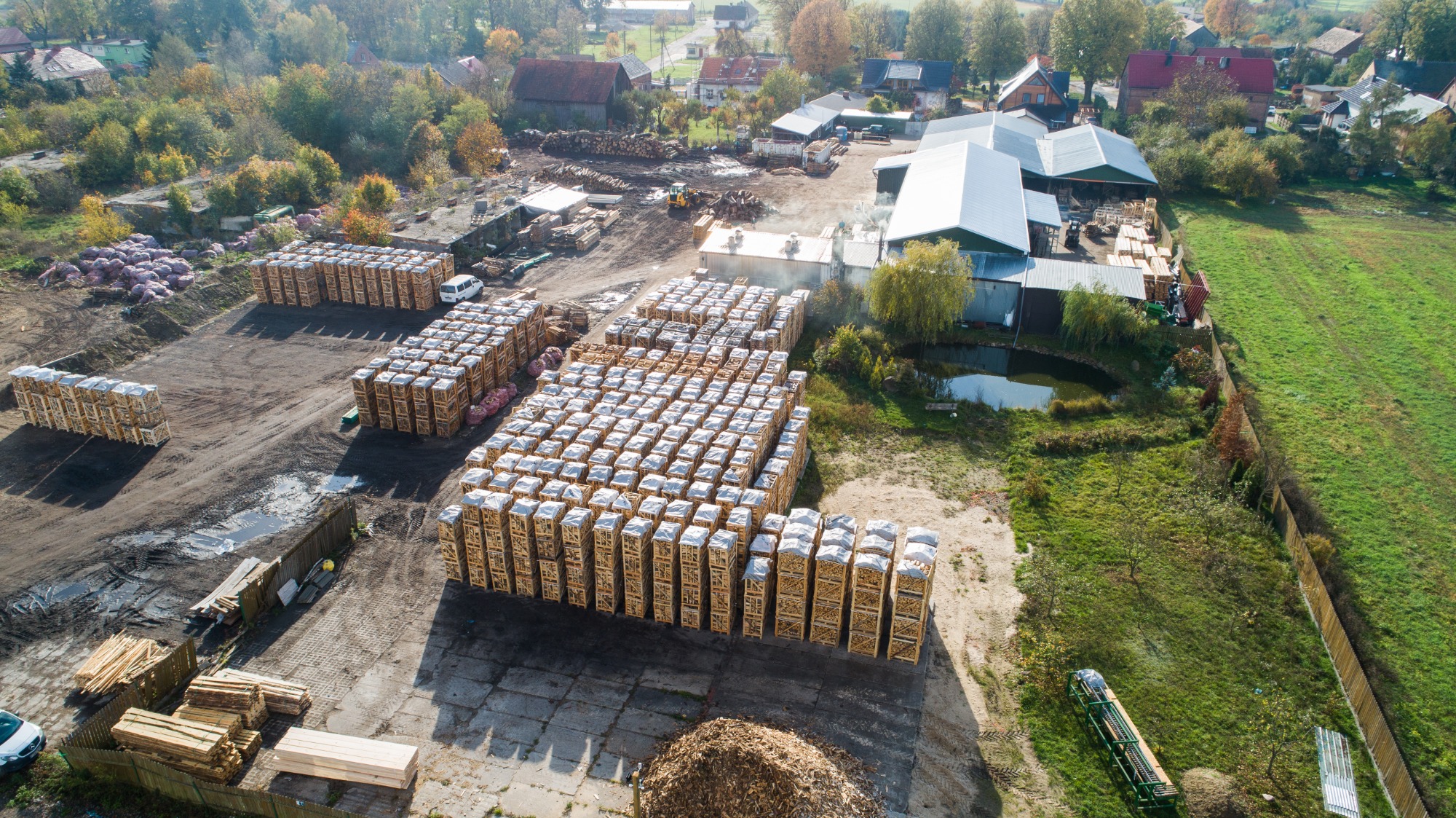 Widok z drona na skład tarcicy w skrzyniach, z budynkami gospodarczymi, stawem i stertą trocin w tle.