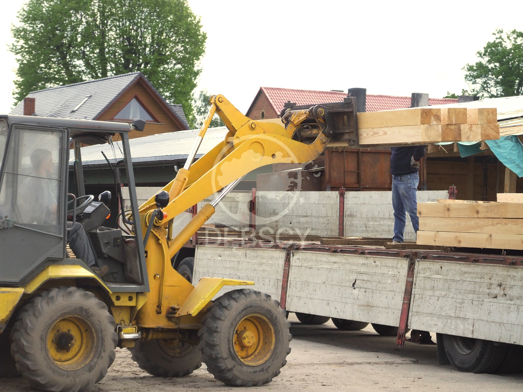 Żółta ładowarka teleskopowa przenosi pakiet świeżych belek na przyczepę ciężarówki; w tle zabudowania gospodarcze.
