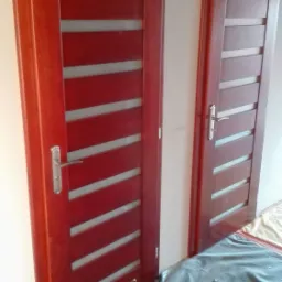 Dwa ciemnoczerwone drzwi wewnętrzne z poziomymi, matowymi przeszkleniami, zamontowane w świeżo pomalowanym pomieszczeniu, zabezpieczonym folią malarską.