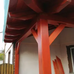 Konstrukcja z belek w kolorze terakoty, prawdopodobnie zadaszenie tarasu, z widocznymi elementami mocującymi i deskami przygotowanymi do montażu przy ścianie budynku.