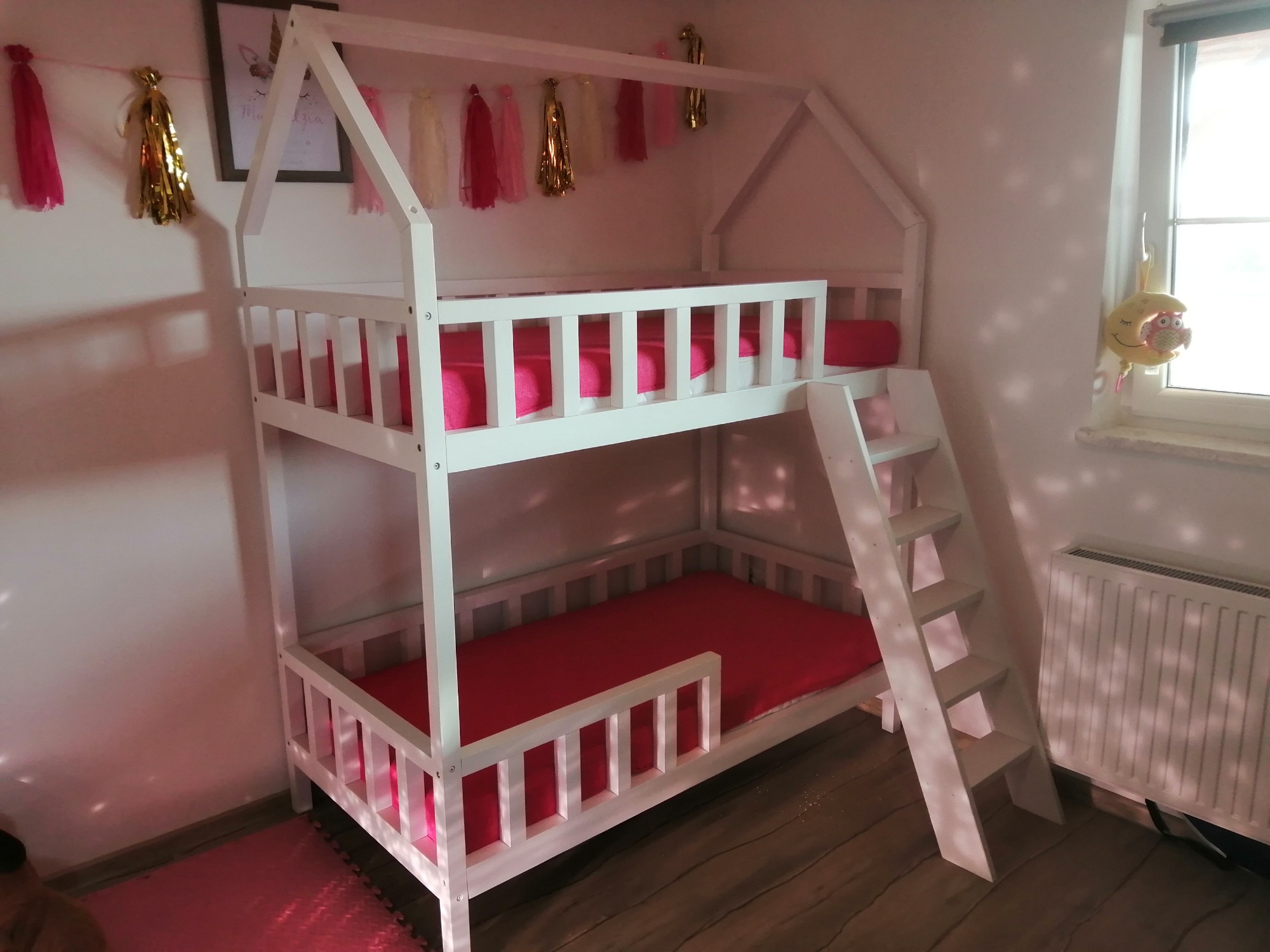 Białe łóżko piętrowe w kształcie domku dla dzieci, z różowymi materacami, dekorowane girlandą z frędzli i obrazkiem na ścianie.