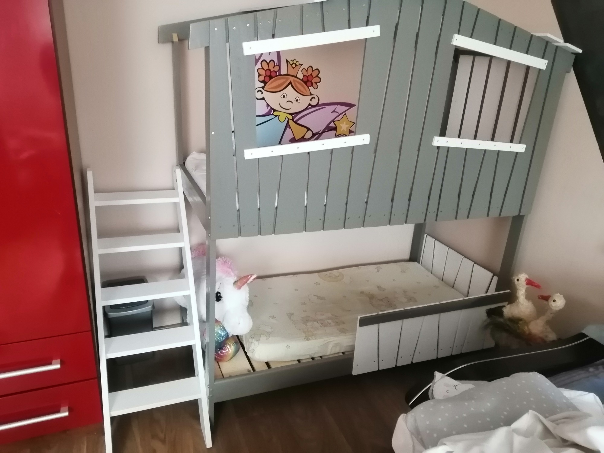 Szare łóżko piętrowe dla dzieci w kształcie domku z drabinką, pluszowym jednorożcem i dekoracją okna z postacią z bajki.