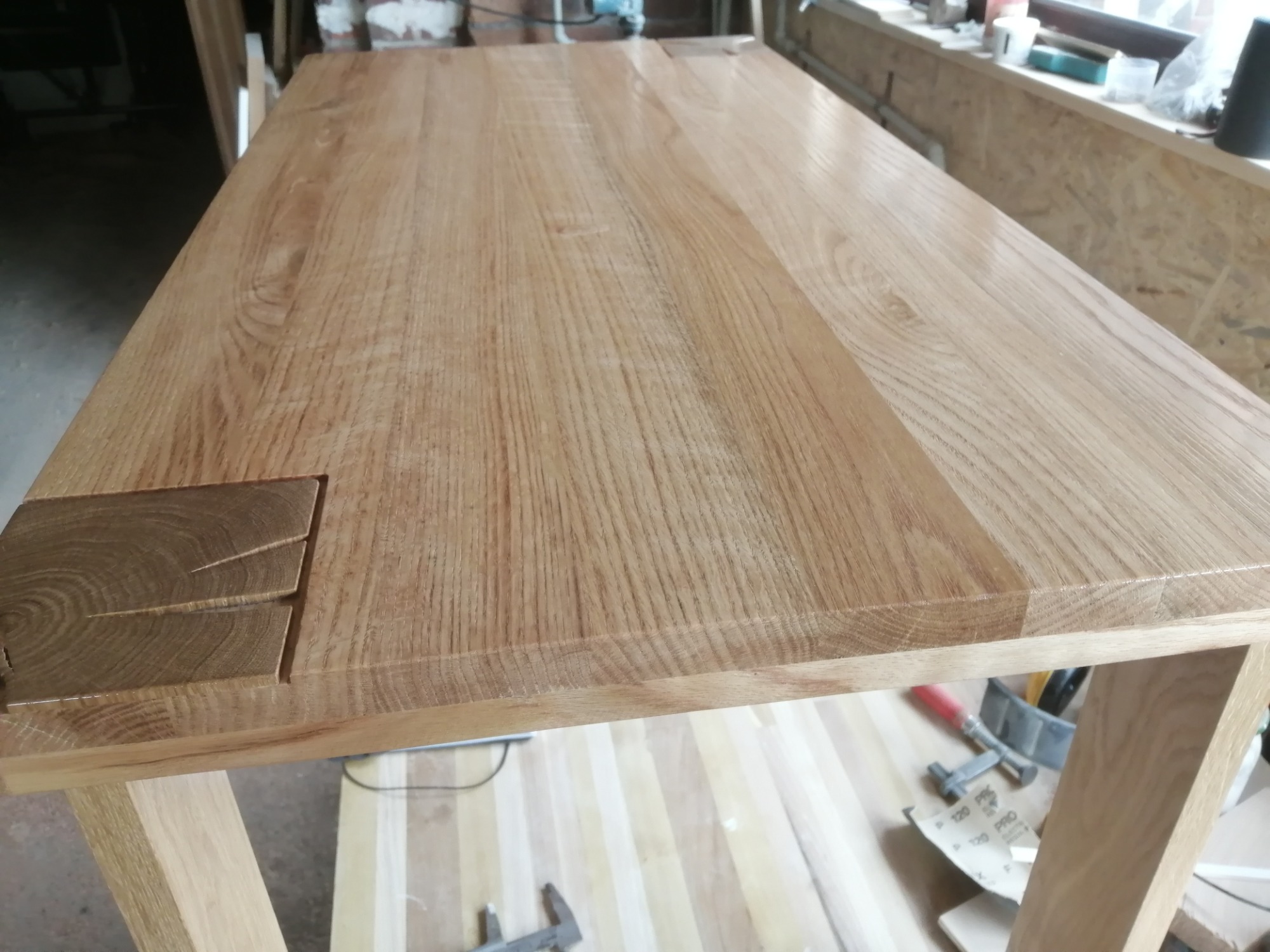 Blat stołu z jasnego drewna dębowego z widocznymi detalami łączenia elementów, w tle warsztat stolarski z narzędziami.